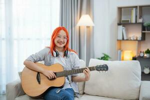 ásia mulher vestindo óculos e fone de ouvido jogando guitarra enquanto sentado em sofá dentro a vivo quarto às lar. ásia mulheres escrevendo música enquanto jogando guitarra às lar. compor música música conceito. foto