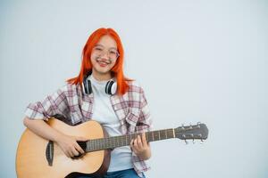 inteligente ásia mulher vermelho cabelo e vestindo óculos músico jogar guitarra em cinzento ou branco fundo. guitarrista música conceito, Ásia mulheres segurando acústico guitarra Veja às a Câmera dentro a estúdio iluminação. foto