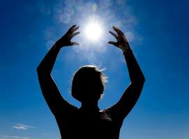 silhueta do uma menina contra a fundo do a Sol e azul céu. mãos estão elevado acima para a Sol foto