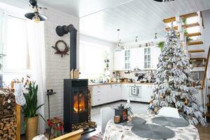 Natal decoração dentro branco cozinha com queimando fogão lareira, festivo bagunça, Vila interior com uma Nevado Natal árvore. Novo ano, Natal humor, acolhedor lar. foto