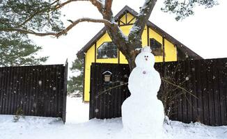 boneco de neve engraçado em um chapéu e cachecol no fundo de uma casa amarela no quintal. inverno, entretenimento de inverno, queda de neve foto