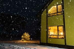 janela festiva aconchegante da casa do lado de fora com a luz quente das guirlandas de luzes de fada dentro - comemore o natal e o ano novo em um lar aconchegante. árvore de natal, bokeh, neve em pinheiros e queda de neve foto