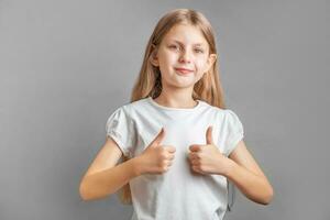 feliz criança, pequeno menina mostrando polegares acima gesto dentro uma branco camiseta foto