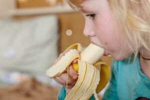 retrato do uma pequeno menina comendo uma banana. a conceito do saudável Comida. uma fresco rápido lanche foto