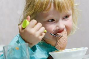 uma pequeno menina tem café da manhã às casa espaguete com salsichas. pequeno Loiras menina comendo jantar com garfo às mesa foto