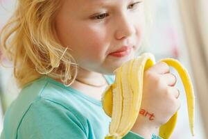 retrato do uma pequeno menina comendo uma banana. a conceito do saudável Comida. uma fresco rápido lanche foto