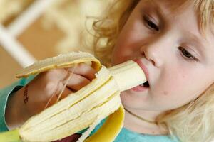 retrato do uma pequeno menina comendo uma banana. a conceito do saudável Comida. uma fresco rápido lanche foto