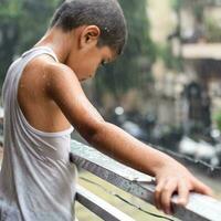 pequeno criança jogando dentro verão chuva dentro casa sacada, indiano inteligente Garoto jogando com chuva gotas durante monção chuvoso temporada, criança jogando dentro chuva foto