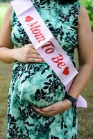 uma grávida indiano senhora poses para ao ar livre gravidez tiro e mãos em barriga, indiano grávida mulher coloca dela mão em dela estômago com uma maternidade vestir às sociedade parque, grávida lado de fora maternidade tiro foto