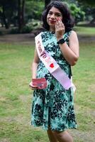 uma grávida indiano senhora poses para ao ar livre gravidez tiro e mãos em barriga, indiano grávida mulher coloca dela mão em dela estômago com uma maternidade vestir às sociedade parque, grávida lado de fora maternidade tiro foto