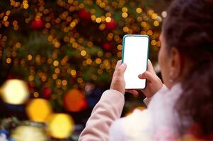 fechar-se mulher mãos usando Móvel telefone, fotografando a iluminado Natal árvore às natal justo, Enviando Móvel aplicativo foto