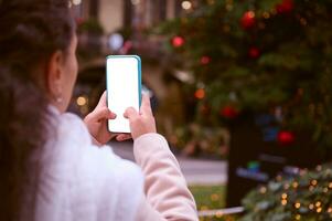 fechar-se mãos do uma mulher segurando inteligente Móvel telefone com branco em branco brincar digital tela, em pé de Natal árvore foto