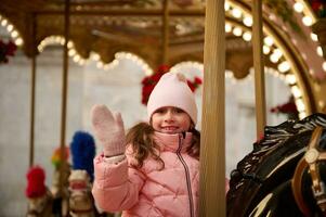 autêntico pequeno menina tendo Boa Tempo equitação uma carrossel cavalo às Natal parque de diversões. conceito do diversão parque, justo foto