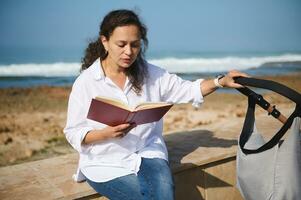 feliz jovem hispânico mulher, mãe empurrando bebê carrinho de bebê, lendo livro, sentado relaxado em a parapeito em atlântico de praia foto