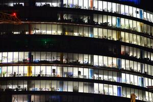 fragmento do a vidro fachada do uma moderno corporativo construção às noite. moderno vidro escritório dentro cidade. grande brilhando janelas dentro moderno escritório edifícios às noite, dentro linhas do janelas luz brilha. foto