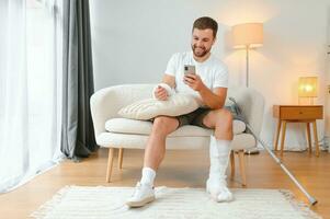 homem com uma quebrado perna e braço usando dele Móvel telefone enquanto relaxante em a sofá às lar. acidente, ferida, tratamento, reabilitação conceito foto