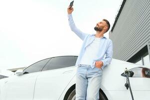 casual homem com Smartphone perto elétrico carro esperando para a terminar do a bateria cobrando processo foto