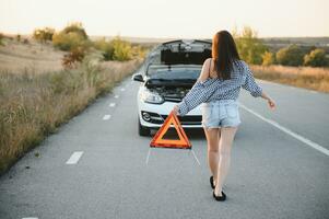 lindo sexy mulher segurando a emergência Pare placa perto uma quebrado carro. foto