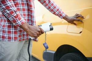 africano homem segurando carregar cabo dentro em mão em pé perto elétrico carro foto