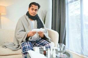 retrato do doente indiano homem com escorrendo nariz segurando papel guardanapos perto face sentado às lar. gripe, vírus, alergia conceito foto