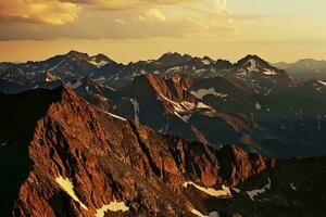 ai gerado uma tirar o fôlego Visão do áspero montanha picos beijou de pôr do sol brilho, adornado com remendos do neve debaixo uma dramático céu. foto