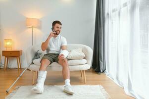 homem com uma quebrado perna e braço usando dele Móvel telefone enquanto relaxante em a sofá às lar. acidente, ferida, tratamento, reabilitação conceito foto