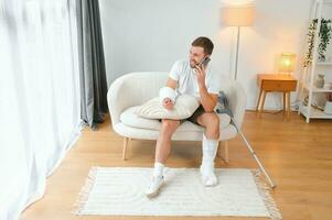 homem com uma quebrado perna e braço usando dele Móvel telefone enquanto relaxante em a sofá às lar. acidente, ferida, tratamento, reabilitação conceito foto