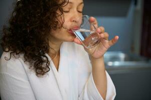 fechar-se encaracolado cabelos bonita mulher fazer trago do água enquanto levando medicamento, uma analgésico ou nutricional suplemento foto