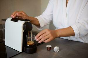 fechar-se do uma jovem mulher dentro branco roupão de banho, fazer dela manhã espresso usando cápsula café máquina para casa usar foto