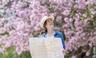 ásia mulher turista segurando cidade mapa enquanto caminhando dentro a parque às cereja Flor árvore durante Primavera sakura flor festival foto