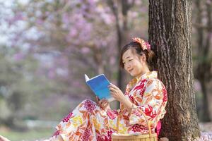 japonês mulher dentro tradicional quimono vestir sentado debaixo cereja Flor árvore enquanto lendo uma livro durante Primavera sakura festival foto