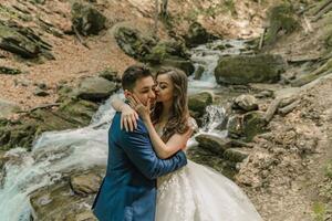 uma Casamento casal abraços de uma montanha rio, ela Beijos ele em a bochecha. noivo e noiva. Casamento foto sessão dentro natureza. foto sessão dentro a floresta do a noiva e noivo.