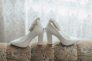 foto do detalhes às a casamento. branco chique sapatos com uma decoração em a pé, em pé em a sofá, perto a janela. contorno linha. sombras e claro.