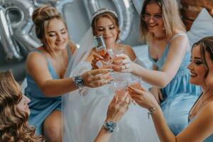 jovem damas de honra dentro azul seda vestidos beber champanhe dentro a da noiva sala. lindo mulheres a comemorar solteira festa sentado em cama e com champanhe. foto