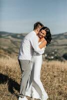 uma à moda modelo casal dentro a montanhas dentro a verão. uma jovem Garoto e uma menina dentro uma branco seda vestir estão caminhando em a declive contra a fundo do a floresta e montanha picos. Garoto Beijos uma menina foto