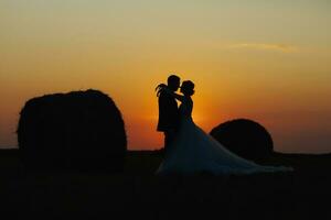 Casamento retrato do a noiva e noivo. noiva e noivo ficar de pé abraçando às pôr do sol perto feno fardos. ruivo noiva dentro uma grandes vestir. à moda, barbudo noivo. silhueta foto. foto