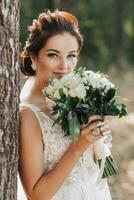 Casamento foto dentro natureza. a noiva é em pé perto uma árvore dentro uma lindo vestir, segurando dela ramalhete do branco rosas, olhando às a Câmera. lindo Maquiagem do a noiva. retrato.