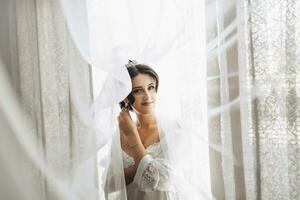 retrato. uma morena noiva é em pé embrulhado dentro uma véu de a janela e olhando abaixo. linda Maquiagem e cabelo. volumoso véu. Casamento foto. lindo noiva foto