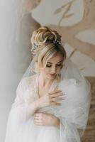 uma noiva com a elegante Penteado, dentro uma manto, carrinhos olhando baixa e segurando dela Branca de Neve véu contra a fundo do pintado paredes. foto