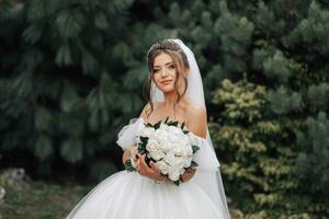 morena noiva dentro uma volumoso branco vestir com aberto ombros e uma grandes véu, sorridente, posando com uma ramalhete do branco rosas. retrato do a noiva. lindo Maquiagem e cabelo. foto