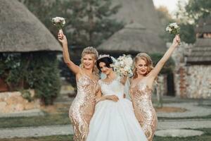 uma morena noiva dentro uma branco elegante vestir com uma coroa e dela Loiras amigos dentro ouro vestidos pose com buquês. Casamento retrato dentro natureza, Casamento foto dentro uma luz tom.