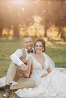 Casamento retrato do a noiva e noivo. feliz recém-casados estão sentado suavemente segurando mãos em a verde grama, posando. à moda noivo. lindo jovem noiva foto