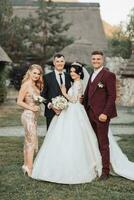 frente Visão do recém-casados caminhando mão dentro mão entre amigos. meninas dentro dourado vestidos aguarde buquês, a noiva dentro uma volumoso vestir, a noivo e dele amigos dentro se adequa. Casamento dentro natureza foto