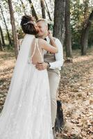 Casamento foto dentro natureza. a noiva abraços e Beijos a noivo. a noiva tem dela ombros virou. lindo Penteado e cabelo decoração. retrato do a noiva e noivo dentro a floresta