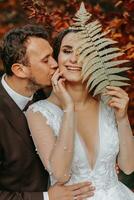 uma menina com uma lindo Penteado é em pé dentro uma branco Casamento vestir dentro a outono floresta contra uma fundo do laranja folhas, segurando uma samambaia ramo dentro dela mãos. dela marido Beijos dela atrás dela foto