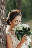 Casamento foto dentro natureza. a noiva é em pé perto uma árvore dentro uma lindo vestir segurando dela ramalhete do branco rosas, olhando às a ramalhete. lindo Maquiagem do a noiva. retrato.