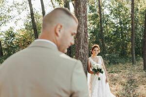 Casamento foto dentro natureza. a noiva é em pé perto uma árvore dentro uma lindo vestir, segurando uma ramalhete do branco rosas, olhando às a Câmera. a noivo é dentro a primeiro plano