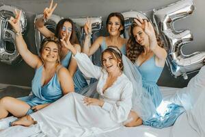 a damas de honra Veja às a sorridente noiva. a noiva e dela alegre amigos estão a comemorar uma solteira festa em a cama dentro idêntico vestidos. noiva e amigos dentro a quarto foto