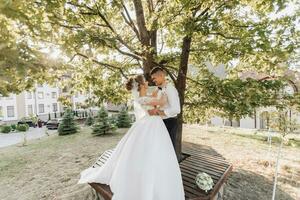 Casamento retrato. uma noivo dentro uma Preto terno e uma Loiras noiva estão de pé, abraçando e se beijando debaixo uma árvore. foto sessão dentro natureza. uma lindo raio do a Sol dentro a foto. lindo cabelo e Maquiagem