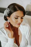 Penteado com a elegante Casamento cabelo acessório. a noiva suavemente toques e mostra a brincos. □ Gentil e à moda Maquiagem foto